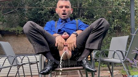 Un uomo in giacca e cravatta con il pene imbracato sta giocando con il suo ano e pisciando in pubblico indossando calzini velati